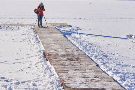 A man shoots video on camera at a pier on a lake in winterの写真素材