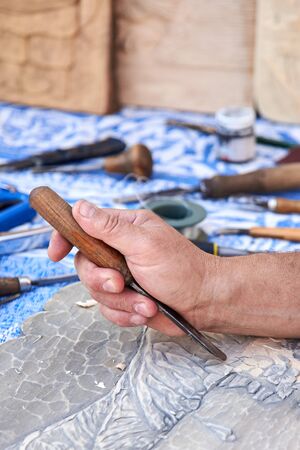 Hands of a man carving a picture of light wood with a cutter.