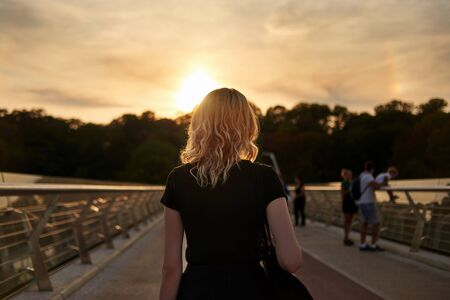 A young blonde woman walks over a city bridge at sunset, back view.