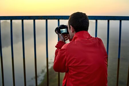 A man on a bridge over the river that photographs a sunrise on a foggy early morning.