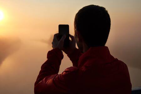 A man on a bridge over the river that photographs on mobile phone a sunrise on a foggy early morning.の写真素材