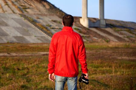 A man in a red jacket with a camera stands near the bridge in the early autumn morning, back view.