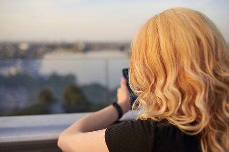 Blonde woman with a mobile phone standing on a bridge over a river, back view.