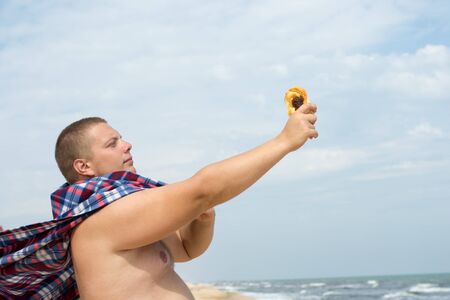 A funny fat man superhero with a plaid instead of a cloak with a bun in hand stands on the sea beach.の写真素材