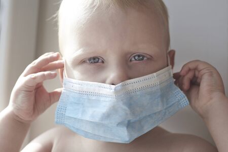 A little blond boy puts a medical mask on his face.