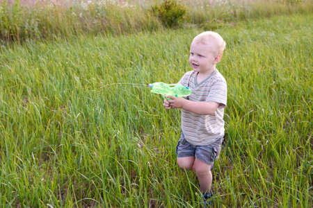 A funny two-year-old boy stands in a meadow with a water pistol, summer fun.の写真素材