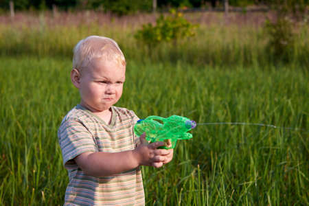 A funny two-year-old boy stands in a meadow with a water pistol, summer fun.の写真素材