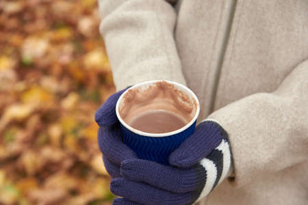 Girl's hands in striped gloves with a cup of hot drink on a blurred autumn background.