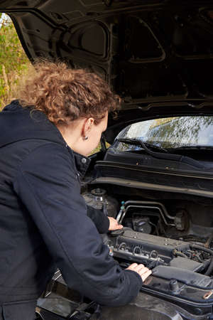 A woman in a jacket is trying to fix a broken car on the road.