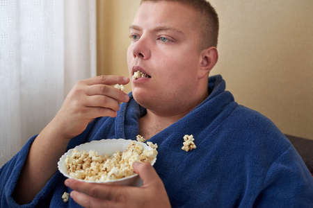 Fat man in a robe eating popcorn while sitting on the couch, obesity and improper lifestyle concept.