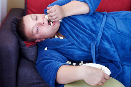 Fat man in a robe eating popcorn while lying on the couch, obesity and improper lifestyle concept.