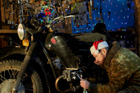 A man in a santa claus hat repairs an old motorcycle in the garage on New Year's Eve.の写真素材