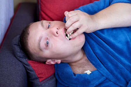 Fat man in a robe eating popcorn while lying on the couch, obesity and improper lifestyle concept.