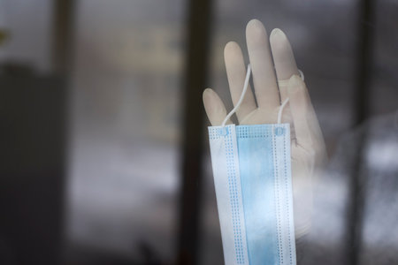 Woman's hand in rubber glove holds a medical mask near the window, self-isolation during the...