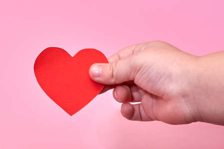 A little boy's hand holds a red paper heart on a pink background, the theme of love and hope.の写真素材