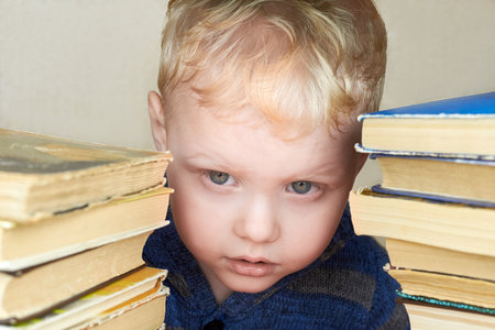 A small three-year-old funny boy looks out from a stack of books.