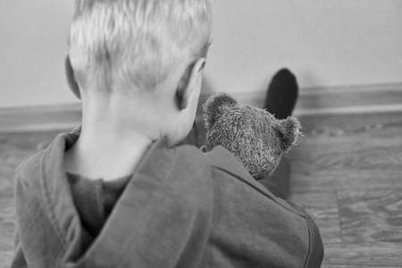 A three-year-old caucasian boy sits alone on the floor and hugs teddy bear, rear view, selective focus, the theme of child abuse, black and white photo.の写真素材