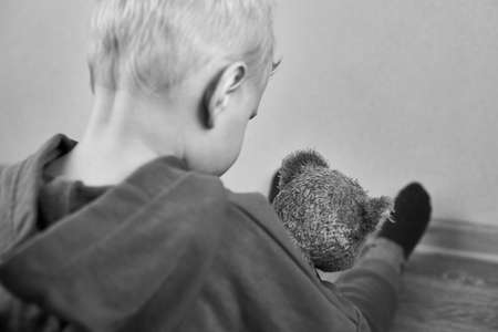 A three-year-old caucasian boy sits alone on the floor and hugs teddy bear, rear view, selective focus, the theme of child abuse, black and white photo.の写真素材