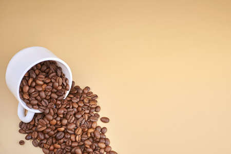 Roasted coffee beans spilling out of a cup on a beige background, copy space.の写真素材