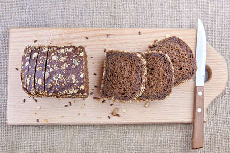 Dark rye sliced bread with whole sunflower seeds, flaxseeds and oatmeal on top on a board with knife, top view.の写真素材