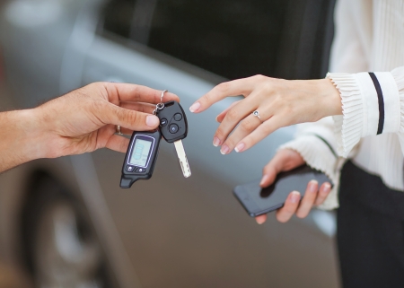 Male hand giving car key to female hand  She is holding a cell phone  In the background, a fragment of the car の写真素材