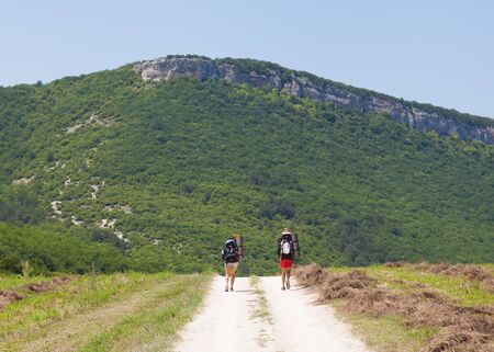 Two tourists with backpacks on the plateau  Young couple go on the road hand in hand  The sun s rays setting sun illuminates them  In the background, mountains covered with forests の写真素材