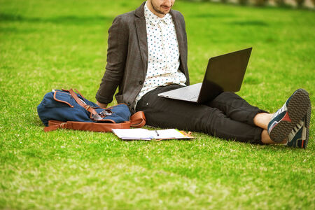 young fashion male student sitting on grass in park and holding a laptopの写真素材