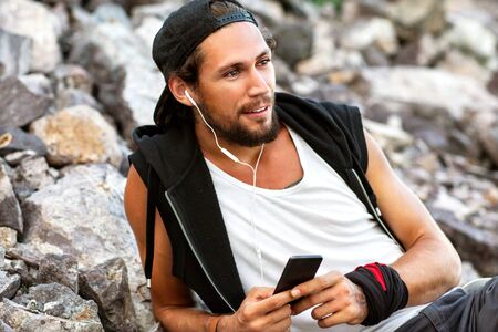 Hipster style boy. Fashion man sitting among the gray stone background is and listening to player