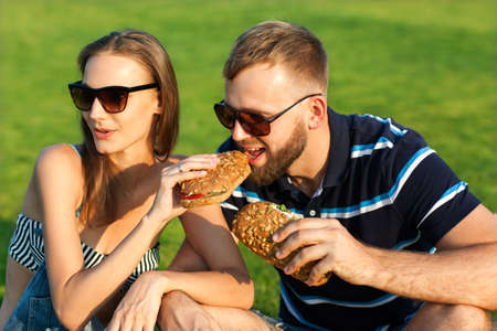couple sitting on the grass in the park and eating sandwiches smilingの写真素材