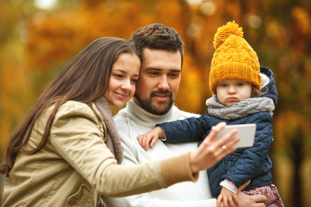 Family with a small daughter happy in autumn park with mobile telephoneの写真素材