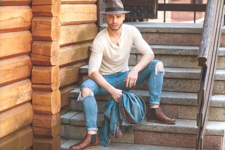 Style fashion hipster guy sitting on the stairs in a light dress and hat on the background of wooden wallの写真素材
