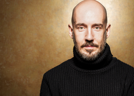 Fashion Portrait of 40-year-old man standing over light gold background in a black sweater. Close up. Classic style. Bald shaved head. Copy-space. Studio shotの写真素材