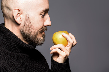 Fashion Portrait of 40-year-old man standing in a black sweater holding an apple. Close up. Classic style. Bald shaved head. Copy-space. Studio shotの写真素材