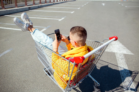 fashion guy in sunglasses and a yellow jacket sits in a cart from food in the supermarket parkingの写真素材