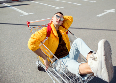 fashion guy in sunglasses and a yellow jacket sits in a cart from food in the supermarket parkingの写真素材