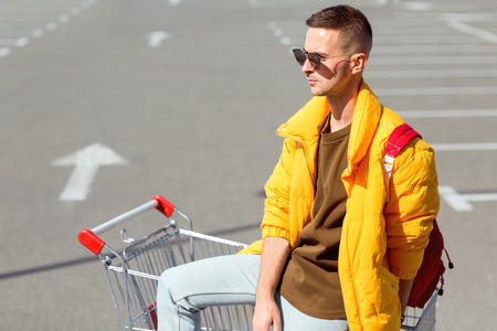 fashion guy in sunglasses and a yellow jacket sits in a cart from food in the supermarket parkingの写真素材