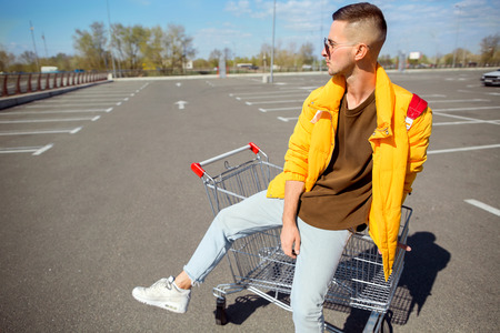 fashion guy in sunglasses and a yellow jacket sits in a cart from food in the supermarket parkingの写真素材