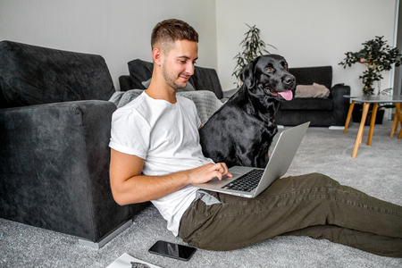 freelancer guy sitting at home working in laptop and with dog in arms, black labrador.の写真素材