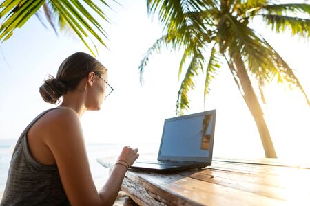 freelancer girl with a computer among tropical palm trees work on the island in sunsetの写真素材