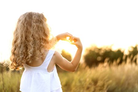 little girl at sunset shows a heart with hands plays in the fieldの写真素材