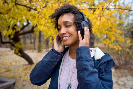 beautiful african girl in autumn park listening to music outdoors smilingの写真素材