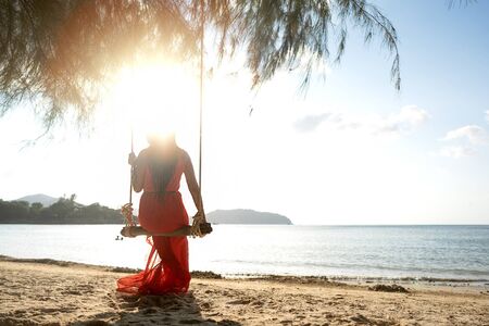 girl in a red dress on a swing among tropical palm trees in Thailand at sunsetの写真素材