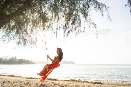 girl in a red dress on a swing among tropical palm trees in Thailand at sunsetの写真素材