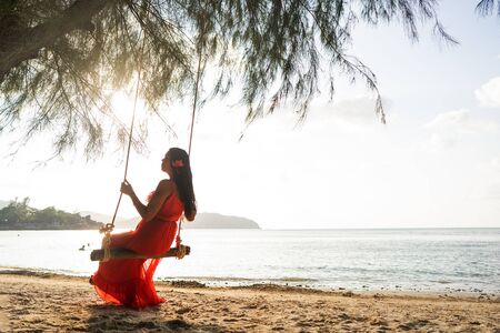 A girl in a red dress on a swing among tropical palm trees in Thailand at sunsetの写真素材