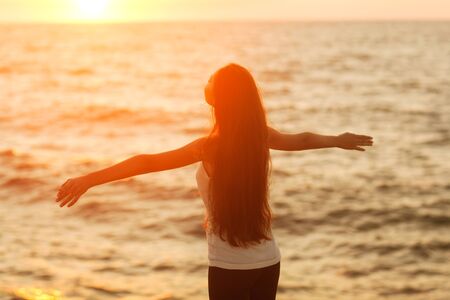 Free woman enjoying freedom feeling happy at beach at sunset. Beautiful serene relaxing woman in pure happiness and elated enjoyment with arms raised outstretched up. Asian Caucasian female model.の写真素材