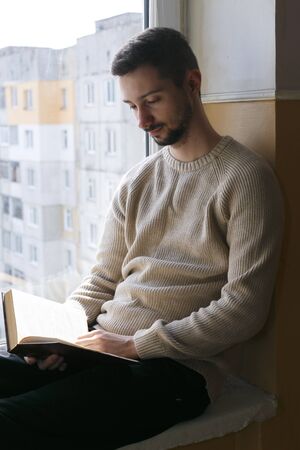 A young man sits on a windowsill and reads a book. A man is wearing a beige sweater. The man is bearded.の写真素材