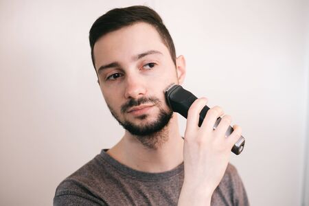 A young man with dark hair shaves his beard. A bearded man mows a beard with a trimmer.の写真素材