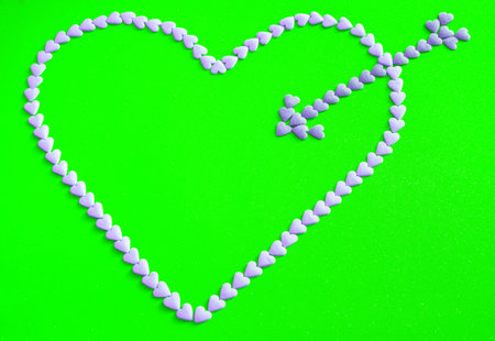 Heart shape pierced by arrow made from litle pink candy hearts on green backgroundの写真素材