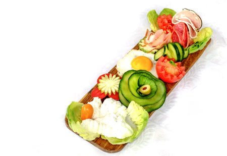 Breakfast on wooden platter - Fresh cheese on lettuce with fried egg, cucumber, tomato, ham and green olives.の写真素材