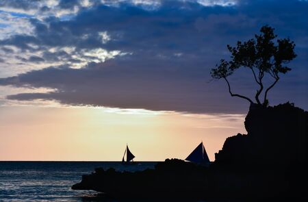 Silhouette of a rock and a tree with some boats in background at sunset in Boracay, Philippinesの写真素材